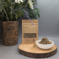 Pouch of dried agrimony herb next to a selenite dish filled with same herb on a wooden plate with a vase and flowers behind.