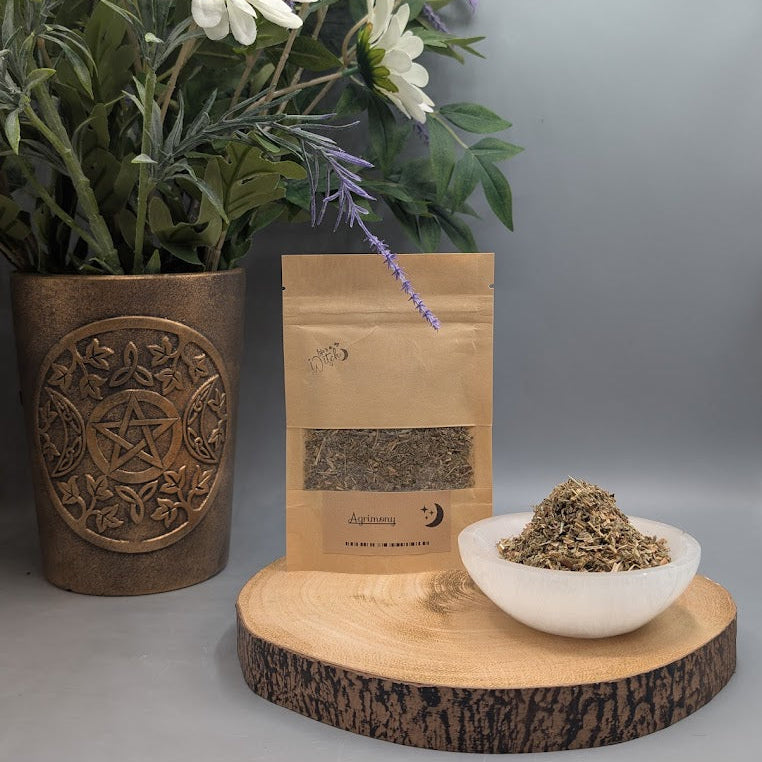 Pouch of dried agrimony herb next to a selenite dish filled with same herb on a wooden plate with a vase and flowers behind.