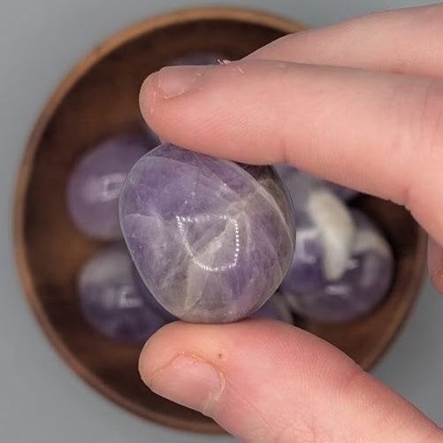 Hand holding a single amethyst tumble with full wooden bowl of the same crystal in background