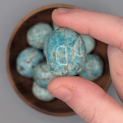 Hand holding a single amazonite tumble with full wooden bowl of the same crystal in background
