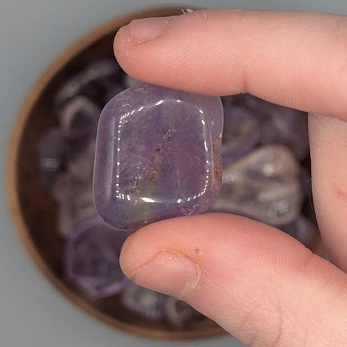 Hand holding single ametrine tumble, over a background of a Brown bowl filled with ametrine tumblestones