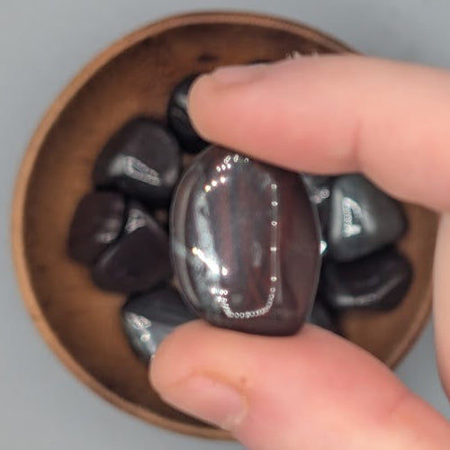Hand holding single ancestralite tumble, over a background of a Brown bowl filled with ancestralite tumblestones