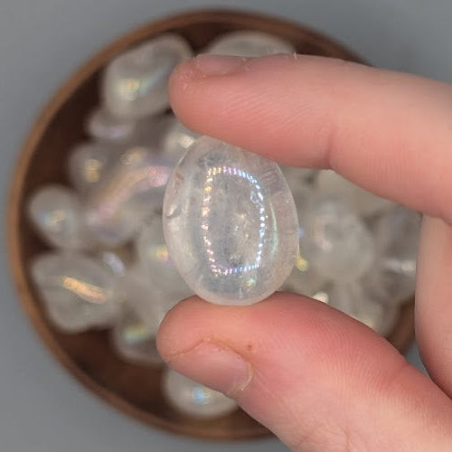 Hand holding single angel aura tumble, over a background of a Brown bowl filled with angel aura quartz tumblestones