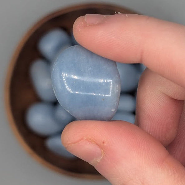 Hand holding a single angelite tumble with full wooden bowl of the same crystal in background