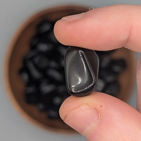 Hand holding a single Apache tear tumble with full wooden bowl of the same crystal in background