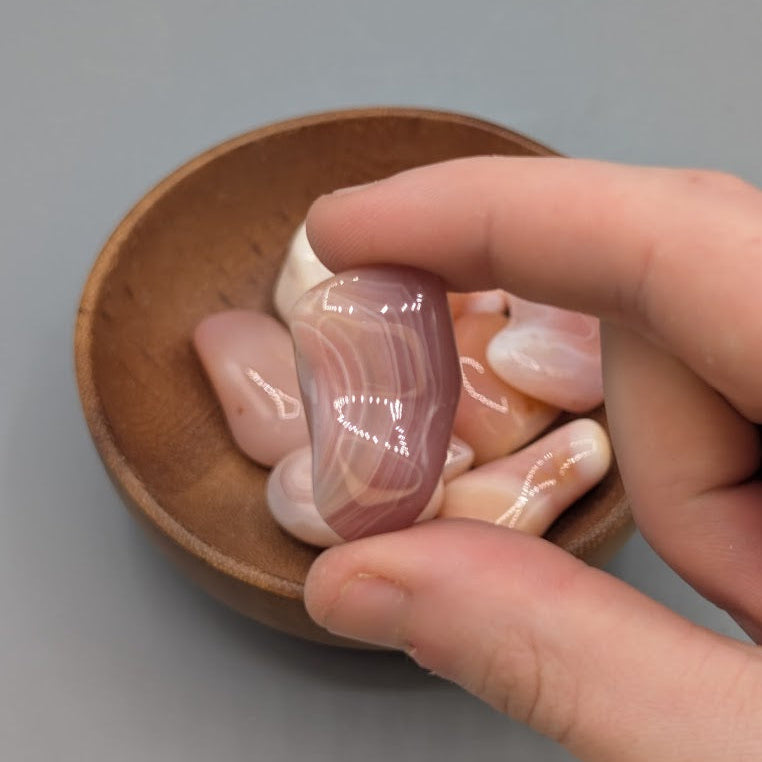 Hand holding a single apricot agate tumble with full wooden bowl of the same crystal in background
