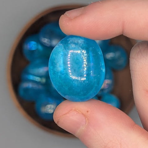 Hand holding a single aqua aura quartz tumble with full wooden bowl of the same crystal in background