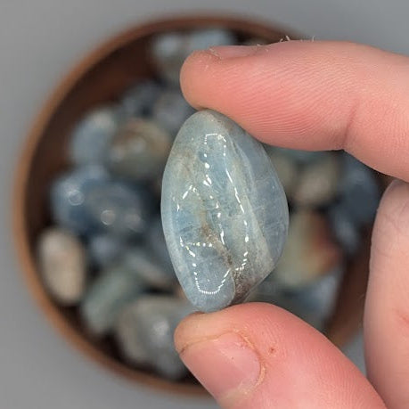 Hand holding a single aquamarine tumble with full wooden bowl of the same crystal in background