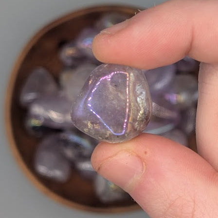 Hand holding a single aura amethyst tumble with full wooden bowl of the same crystal in background