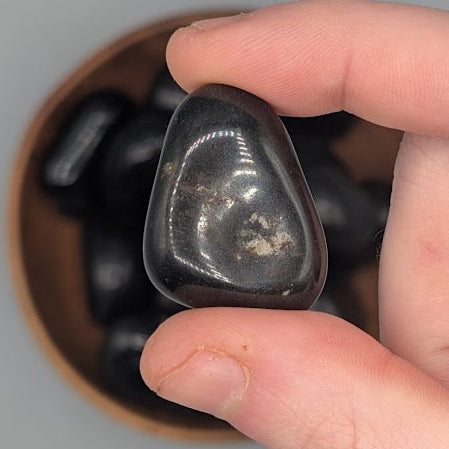 Hand holding a single black agate tumble with full wooden bowl of the same crystal in background