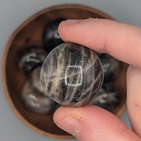 Hand holding a single black moonstone tumble with full wooden bowl of the same crystal in background