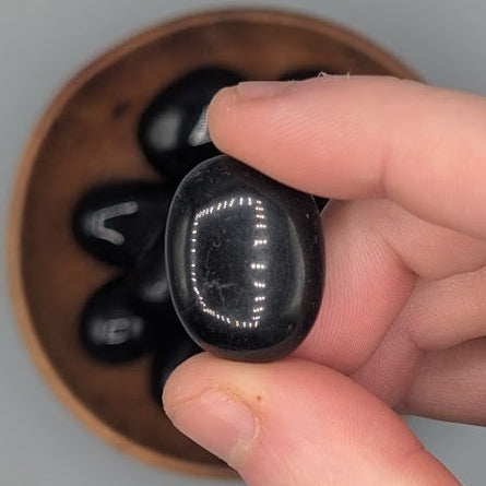 Hand holding a single black obsidian tumble with full wooden bowl of the same crystal in background