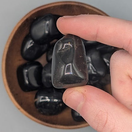 Hand holding a single black onyx tumble with full wooden bowl of the same crystal in background