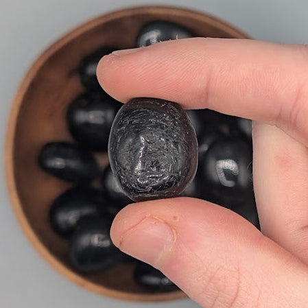 Hand holding a single black tourmaline tumble with full wooden bowl of the same crystal in background