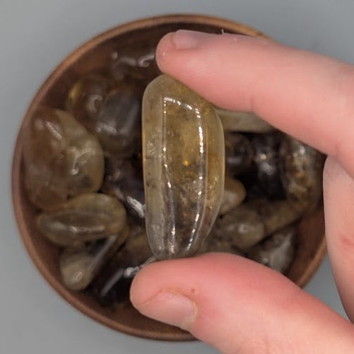 Hand holding single citrine tumble, over a background of a Brown bowl filled with natural citrine tumblestones