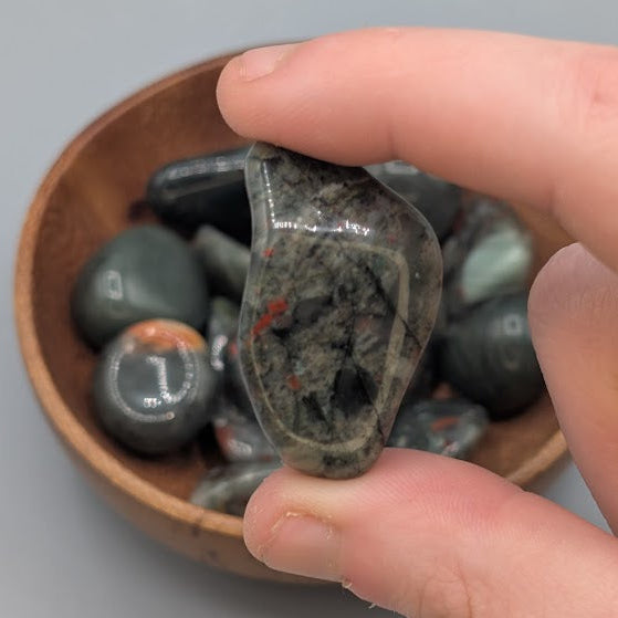 Hand holding a single african bloodstone tumble with full wooden bowl of the same crystal in background